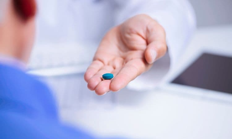 A doctor in a white coat holding out a single blue tablet on an open palm to a seated patient