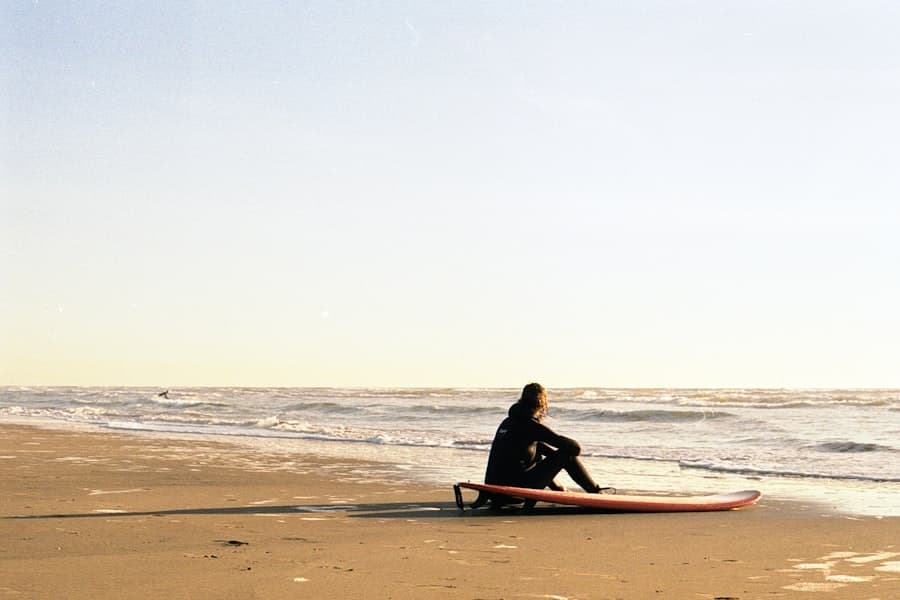 A person in a wetsuit sitting beside a red surfboard on an empty beach at sunset