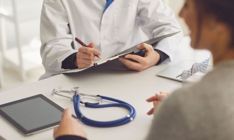 A doctor in a white coat writing on a clipboard across a desk from a patient, a stethoscope and tablet beside them