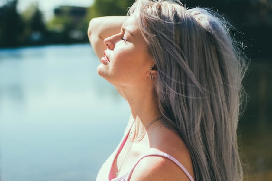 A woman with long grey-blonde hair tilting her head back in the sunlight beside a lake