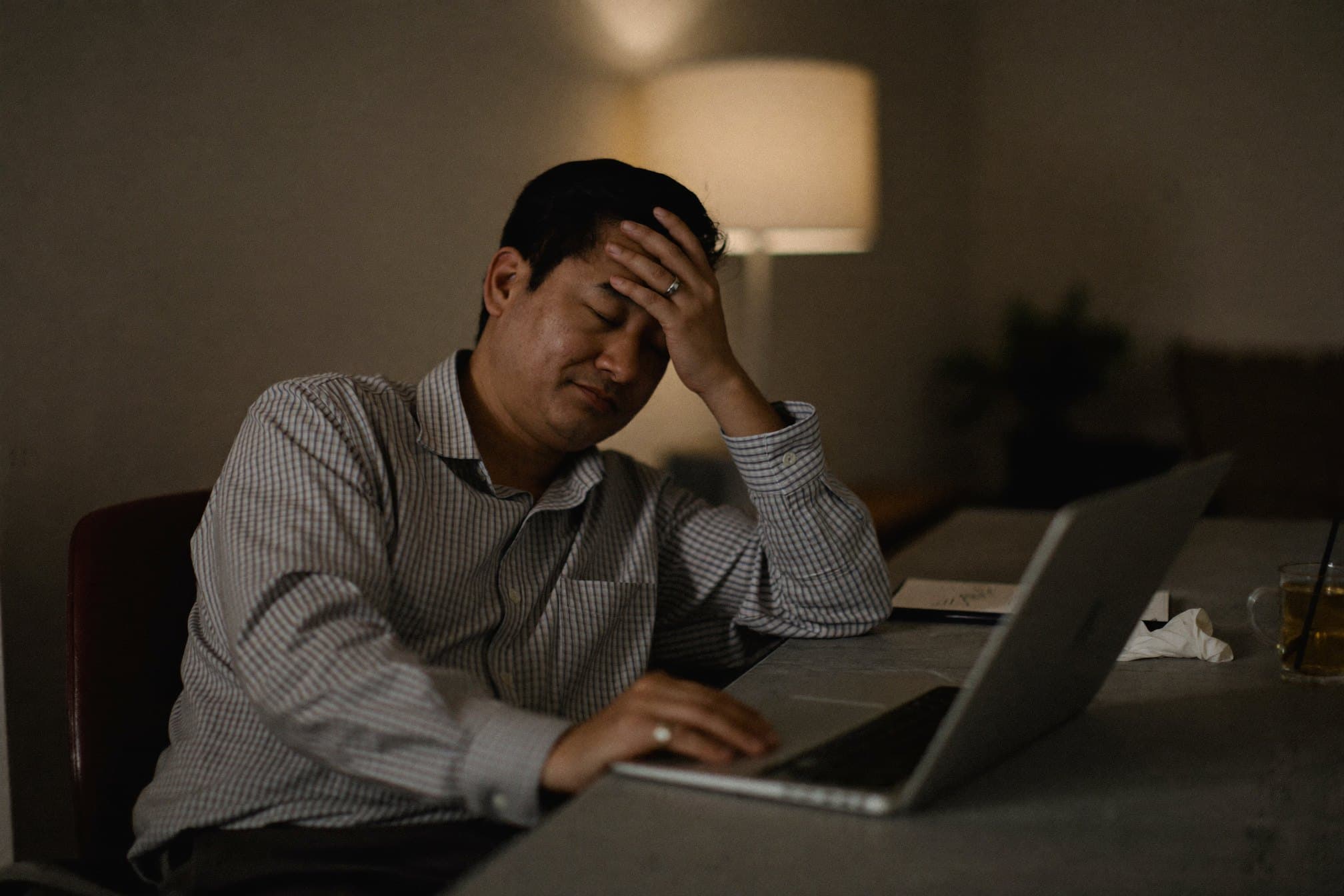 A tired man at his laptop late in the evening, hand on his forehead