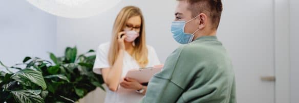 一名戴口罩的患者在诊症室与医护人员交谈，对方正在写字板上做记录。