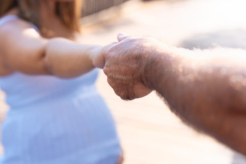 A pregnant woman in a white dress holding her partner's hand outdoors in bright sunlight
