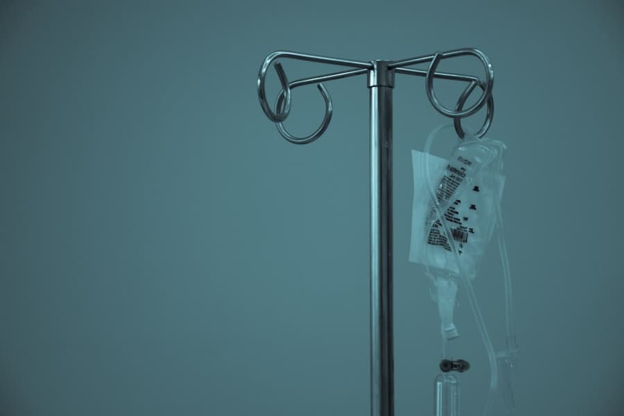 A metal IV drip stand holding a saline bag and tubing against a plain teal wall