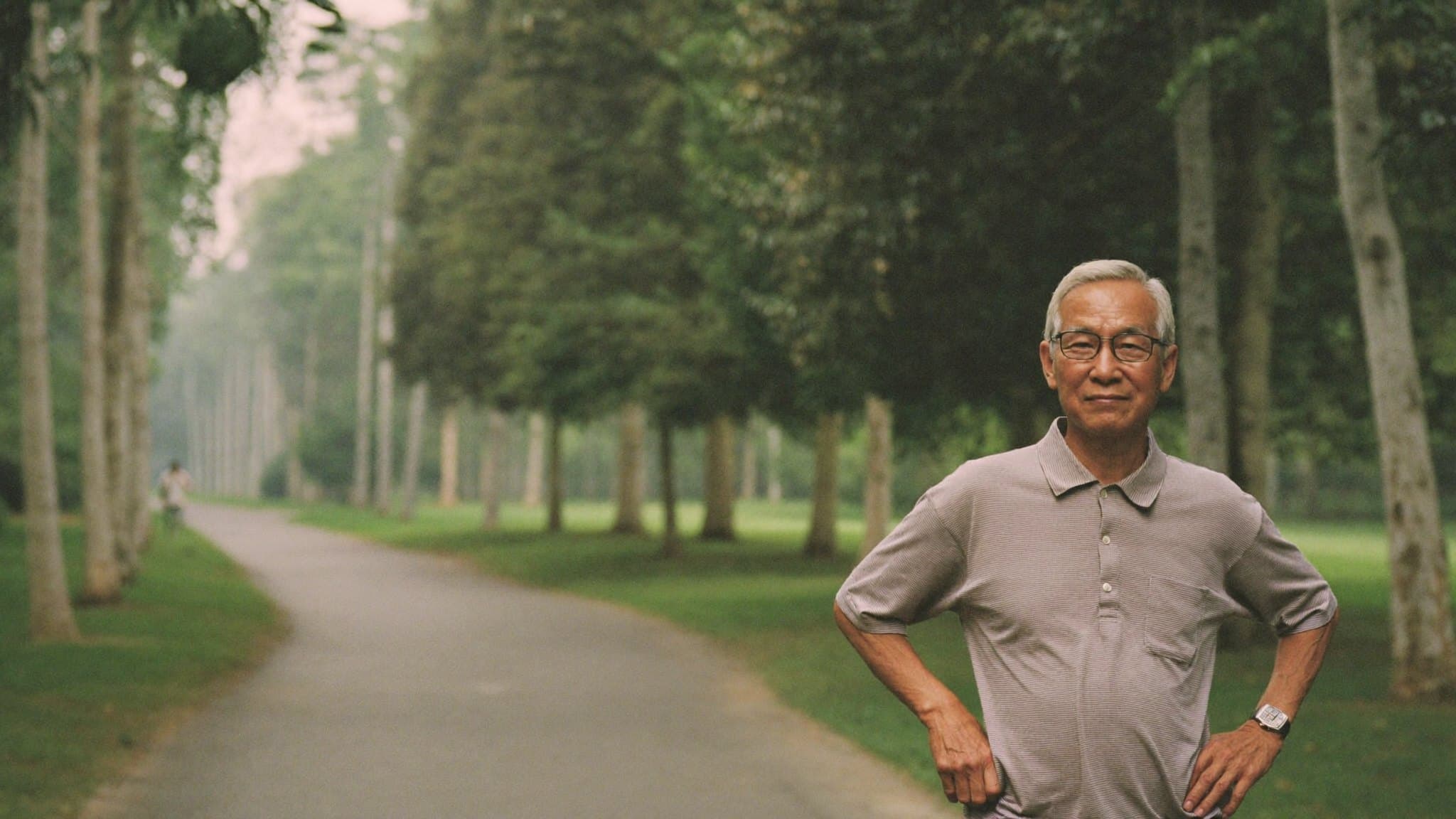 Seorang lelaki berusia berdiri di laluan taman yang dipenuhi pokok pada awal pagi, tangan di pinggang.